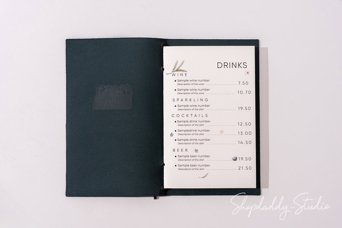 A dark teal or black menu book with an open page showing a printed drinks list, photographed from above on a plain white background.