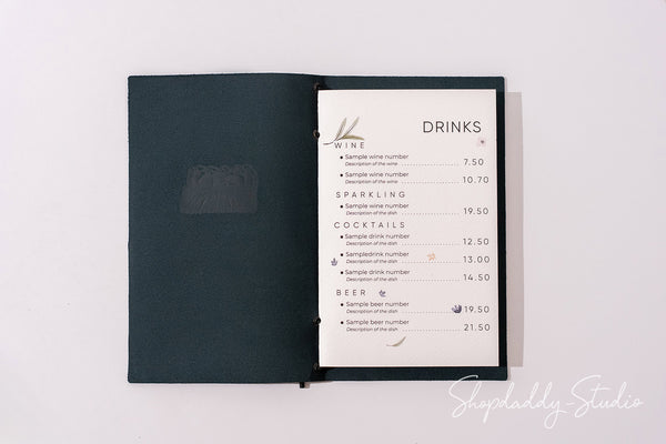 A dark teal or black menu book with an open page showing a printed drinks list, photographed from above on a plain white background.