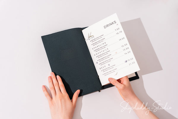 A top-down view of a person's hands holding an open black hardcover journal or menu, with a white printed 'DRINKS' page partially pulled out. The background is a clean white surface with soft shadows.
