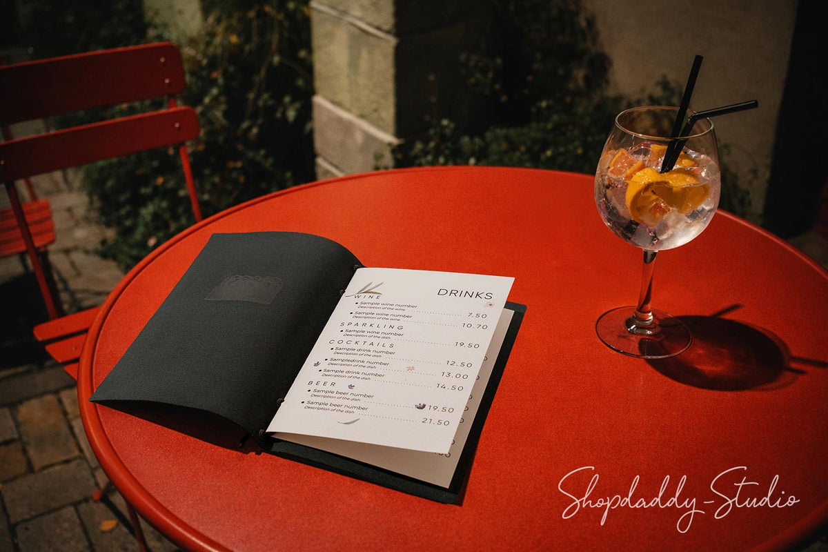 An open black menu on a round red outdoor table next to a glass of cocktail with orange slices and a black straw, with a red chair and stone wall in the background.