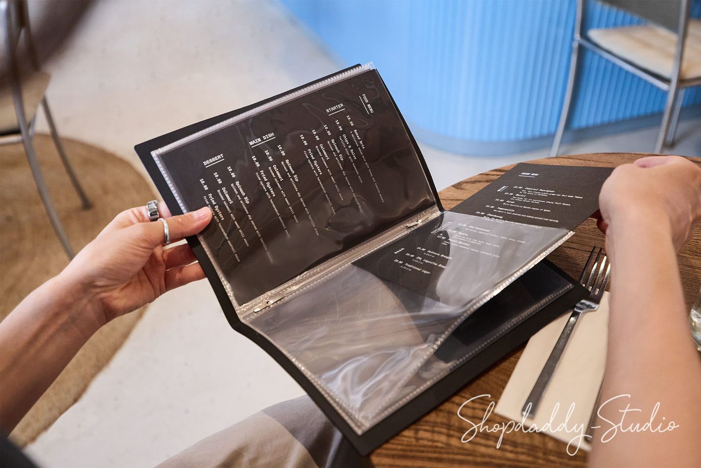 A person holding an open black paper menu in a restaurant setting, with a fork and knife on a wooden table in the background.