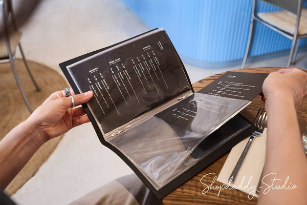 A person holding an open black paper menu in a restaurant setting, with a fork and knife on a wooden table in the background.