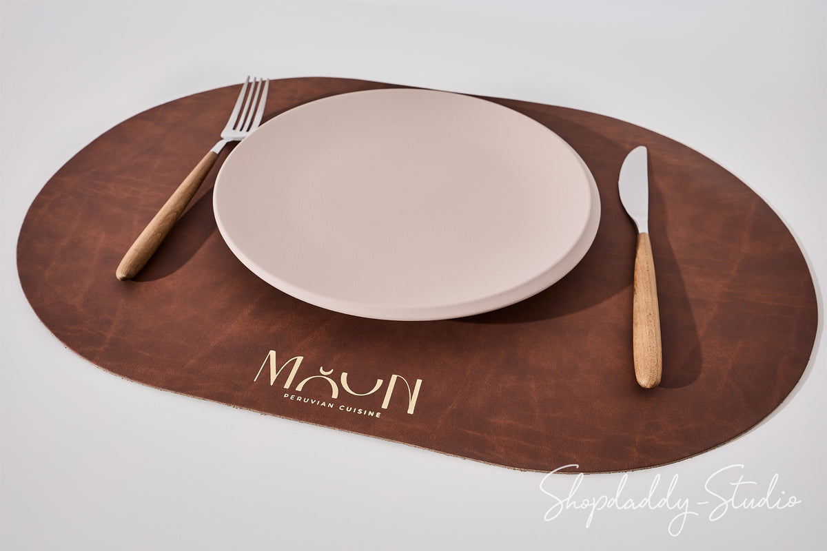 An oval brown leather placemat on a light gray background, styled with a beige ceramic plate, a wooden-handled fork on the left, and a matching knife on the right. The placemat features the brand name 'MOON' printed in gold near the bottom.