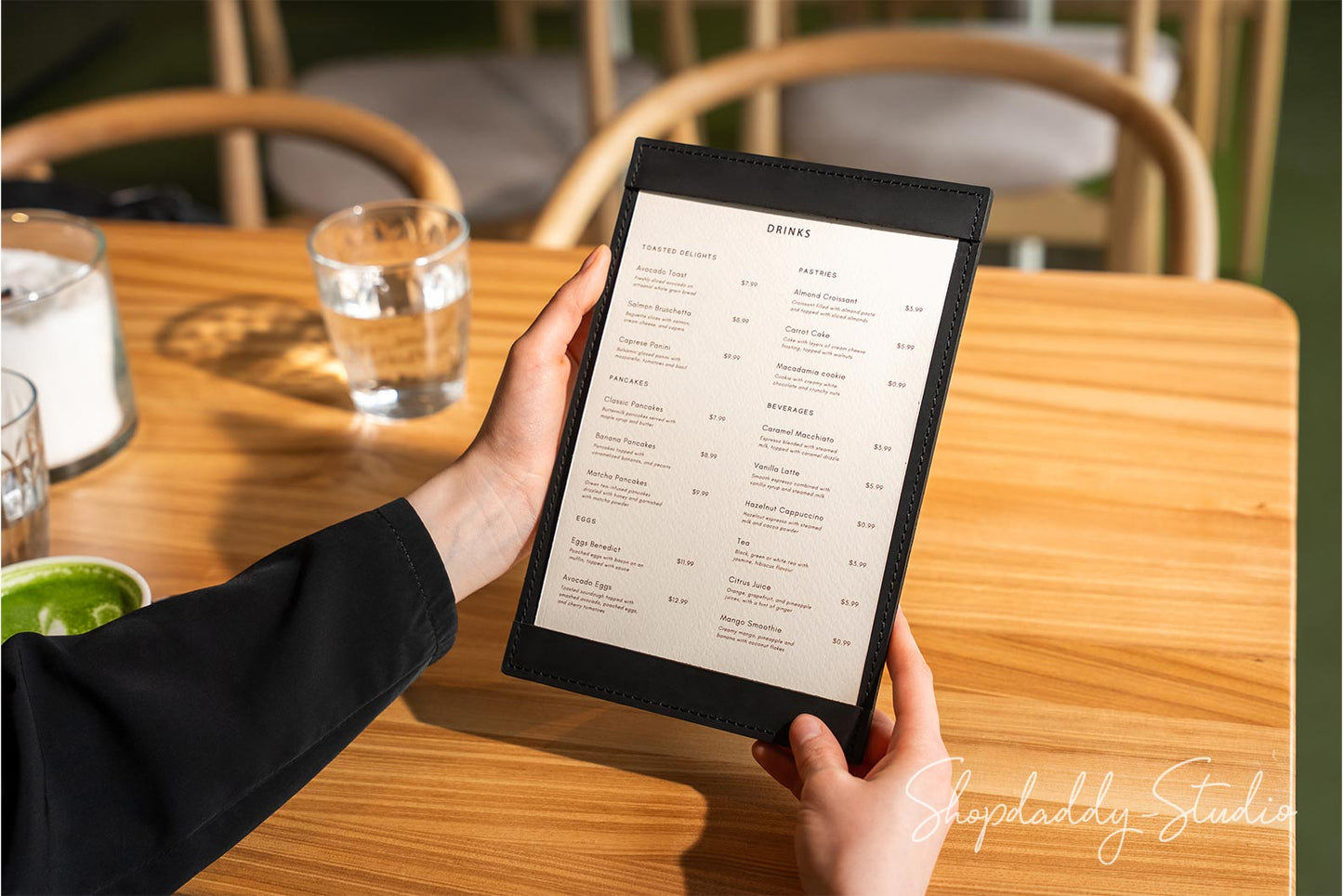 A person holding a black leather-bound paper menu at a wooden table in a restaurant setting, with chairs and drinks blurred in the background.