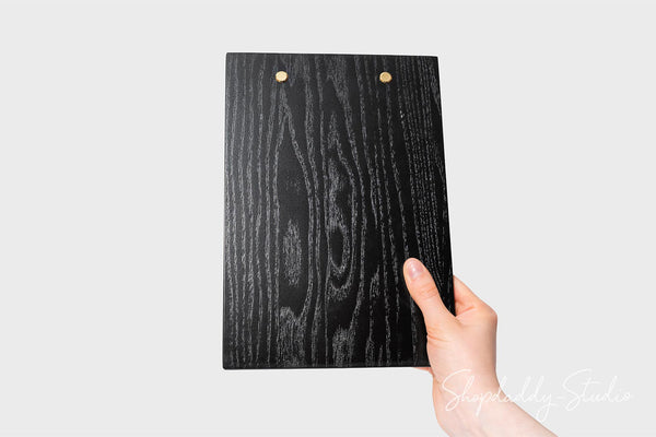 A hand holding a black clipboard-style board with a dark woodgrain pattern and two small gold metal rivets, shown against a light gray background.