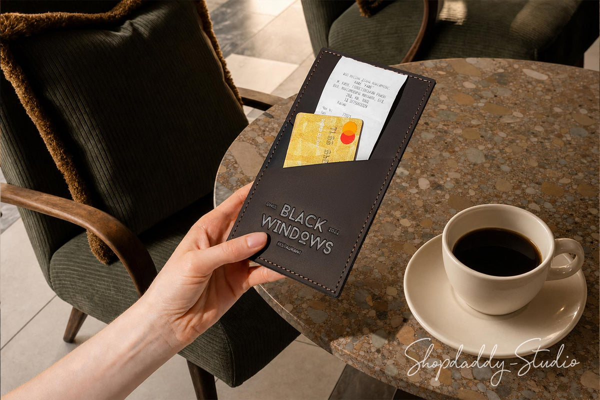 A hand holds a black leather card holder pouch branded 'BLACK WINDOWS' in a cafe setting with a coffee cup on a table in the background.