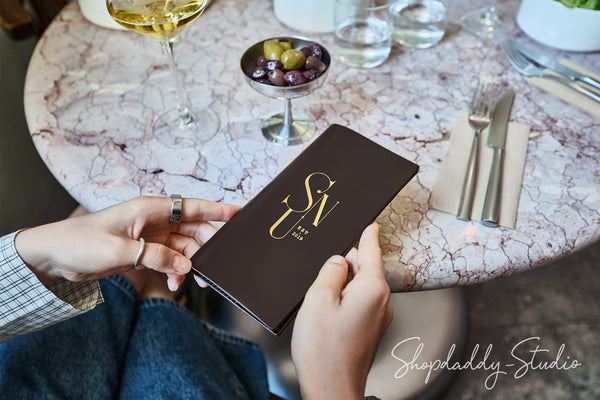 A person holds a dark brown leather menu or notebook with gold lettering reading 'SVL EST. 2024' over a round pink marble restaurant table with wine, olives, and cutlery.