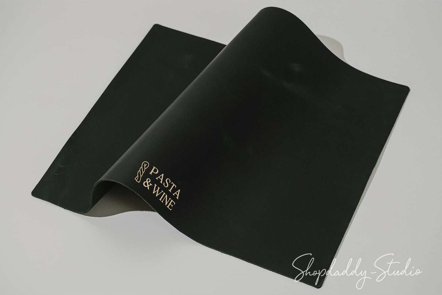 A green rectangular placemat with gold foil lettering, shown partially unrolled on a light gray background.