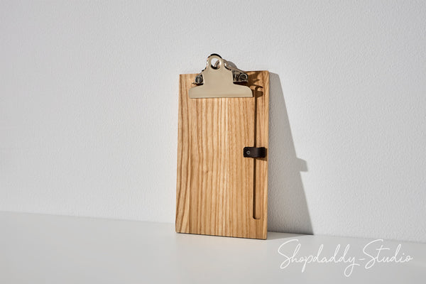 A small wooden clipboard with a light natural wood grain finish, a silver metal clip, and a small black latch, standing upright against a white wall.