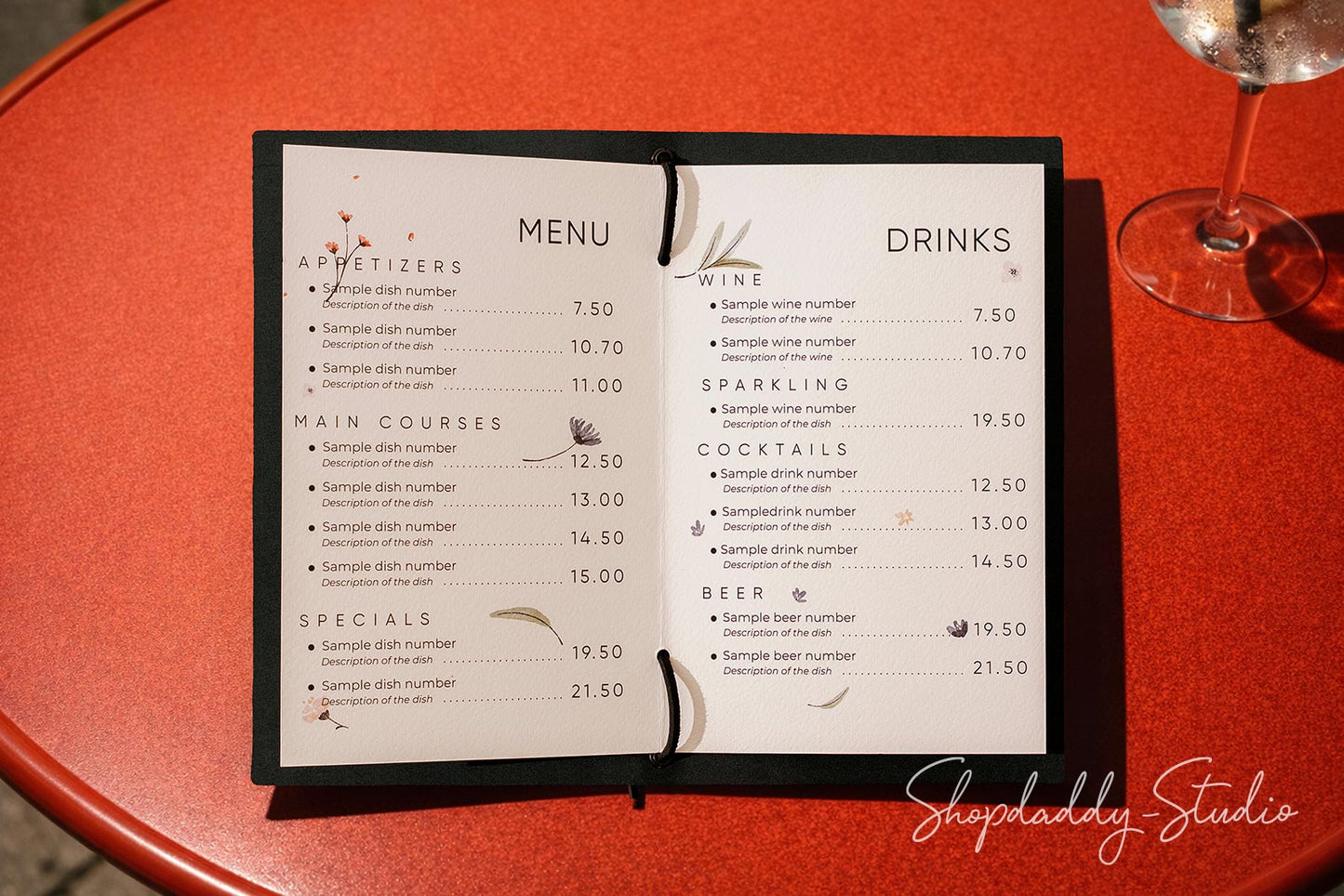 An open black-framed printed menu with appetizers, main courses, specials, wine, sparkling, beer, and drinks listed on white pages, placed on a red table with part of a wine glass visible at the upper right.
