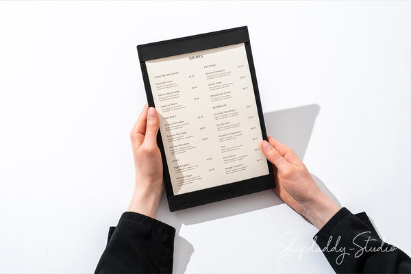 A top-down view of a person holding a black-framed menu board with a cream-colored printed page. The person's hands and black sleeves are visible against a clean white background with a soft shadow.