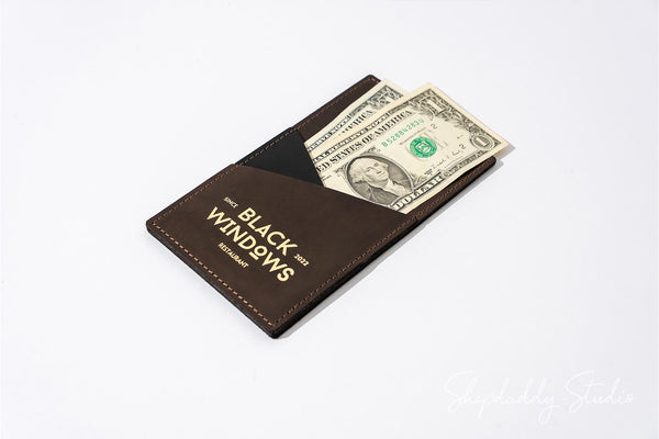 A dark brown leather money clip wallet with gold lettering that reads 'BLACK WINDOWS' and 'BETTER WINDOWS TOGETHER', holding a US dollar bill, shown on a white background.