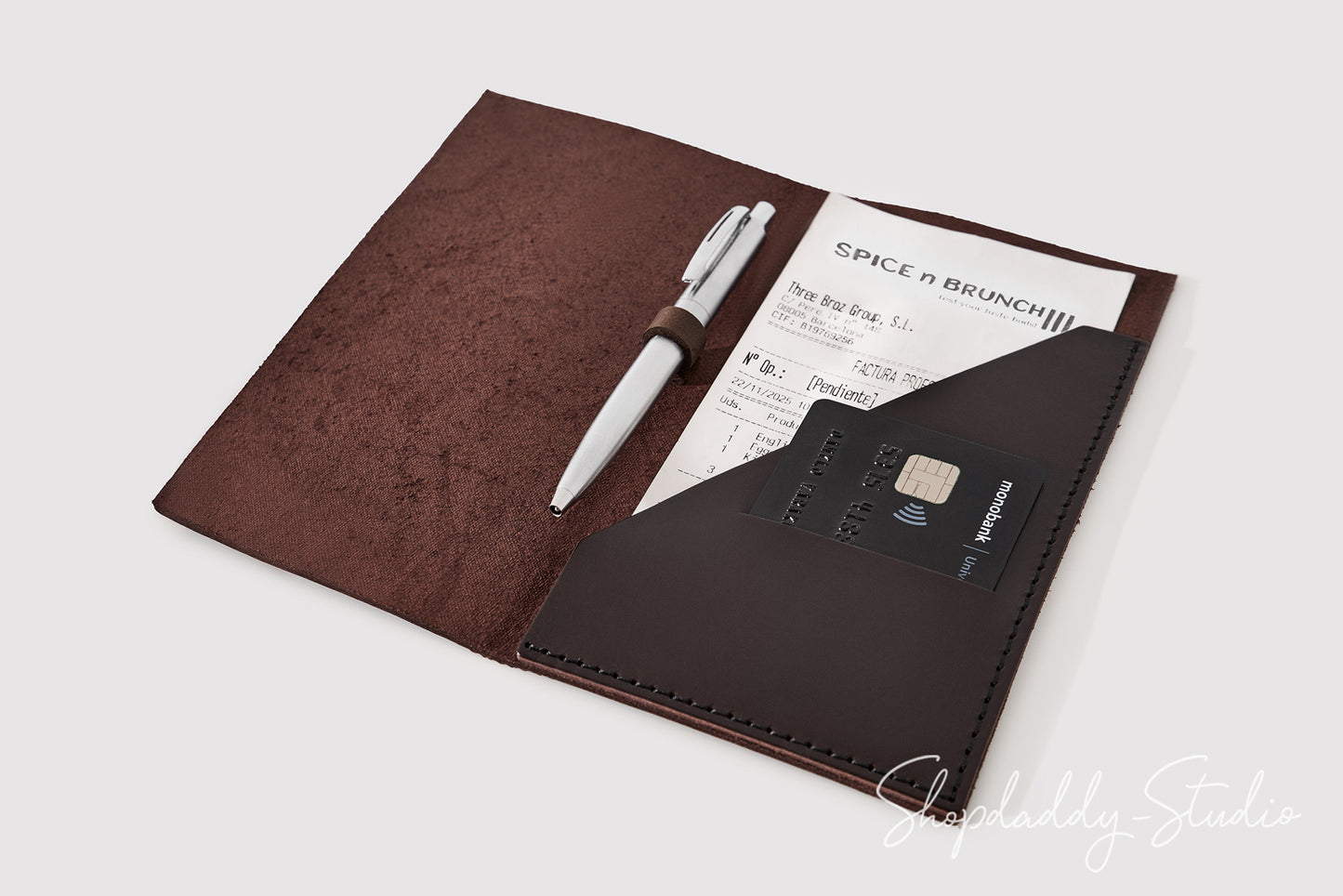 A brown faux-leather travel pass holder or organizer shown open on a light gray background, containing a white pen, a printed paper ticket, and a black credit card in a pocket.