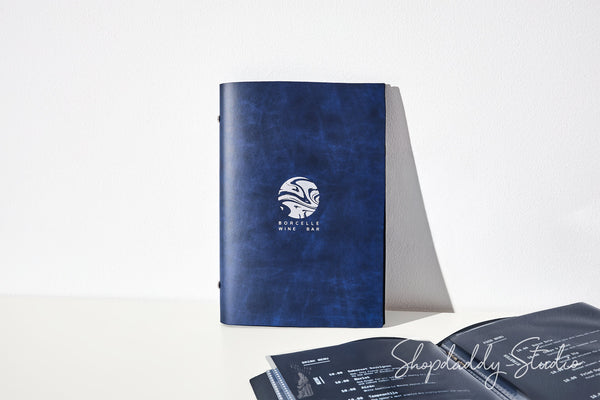 A dark blue textured leather-bound notebook with a white circular logo stands upright on a white surface, with an open booklet spread out in the foreground. The background is a clean white studio setting.