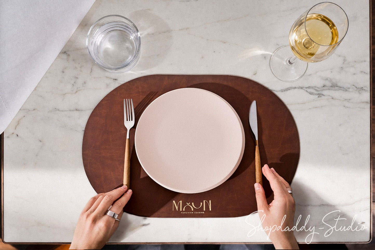 Top-down view of a place setting on a white marble table, featuring a dark brown oval placemat with the name 'MAYUN' printed in gold at the bottom, a white ceramic plate, a fork with a gold handle, a knife with a gold handle, and two hands holding the cutlery. A glass of white wine and a glass of water are also visible.