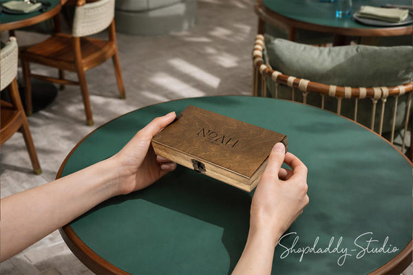 A person holds a small rectangular wooden box with a clasp closure over a round green table in a cafe setting. The box is engraved with the name 'NOAH'.