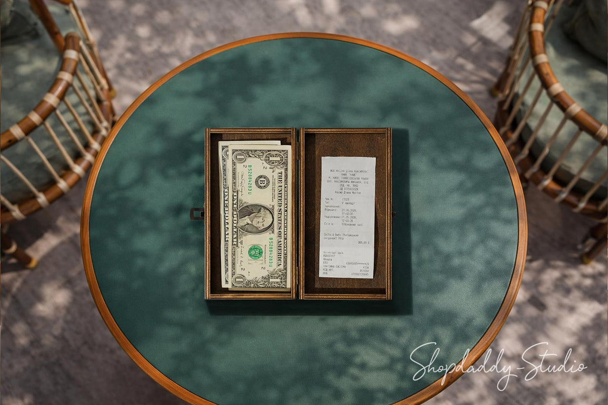 An overhead shot of a round teal-green table with a wooden rim, featuring an open dark wood box containing a stack of one hundred dollar bills and a handwritten receipt. Two woven rattan chairs are partially visible in the background on a gray patterned floor.