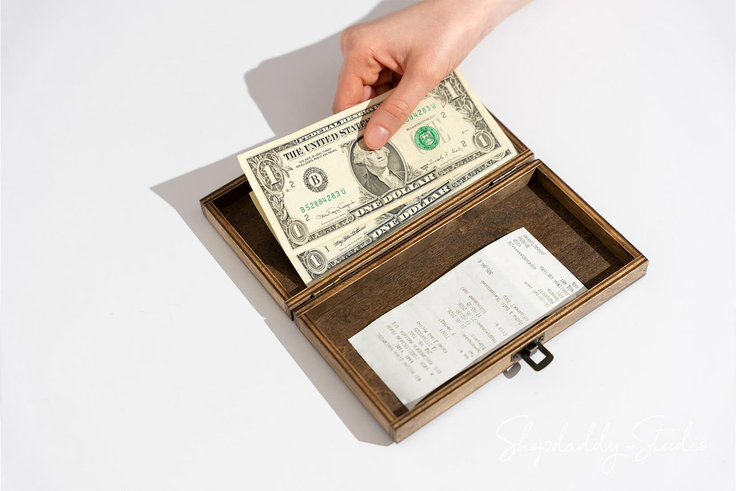 A wooden money box with a brown velvet interior, shown open with a hand placing a one-dollar bill inside and a receipt resting below, on a clean white background.