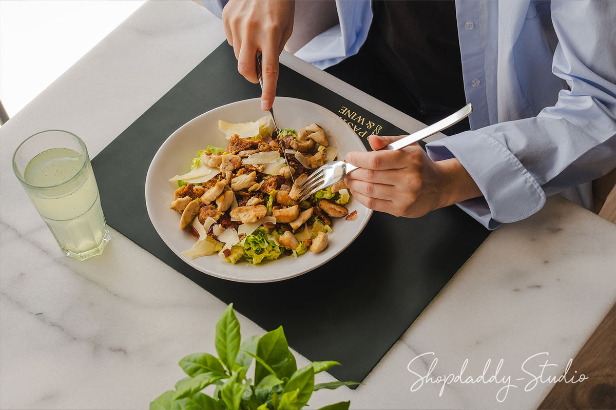A top-down view of a person cutting a salad with a fork and knife on a white plate, placed on a black placemat on a white marble table. A glass of water and green basil leaves are also visible.